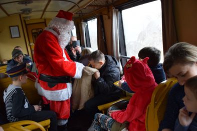 Wagon kolejowy wypełniony jest pasażerami, wśród których dominują dzieci. Osoba przebrana za Świętego Mikołaja rozmawia z dziećmi i wręcza im prezenty. Wszyscy zdają się być w dobrych humorach, a wagon ma żółte obicie foteli i drewniane wykończenia.