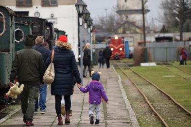 Ludzie spacerują po peronie kolejowym; dorośli trzymają dzieci za rękę. W tle widać zieloną lokomotywę oraz czerwony pojazd szynowy stojący na torach. Na zewnątrz jest dzień i jest pochmurno, a osoby na zdjęciu są ubrane w sezonowe kurtki.