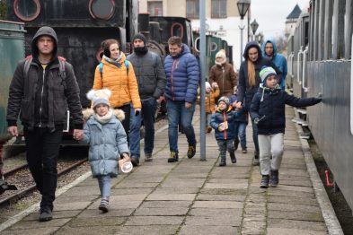 Grupa ludzi, w tym dorosłych i dzieci, spaceruje wzdłuż parkującego pociągu wąskotorowego. Osoby są ubrane w zimowe kurtki i szaliki, co sugeruje chłodną pogodę. Niebo jest zachmurzone, a tory kolejowe oraz pociąg wskazują na stację lub muzeum kolejnictwa.