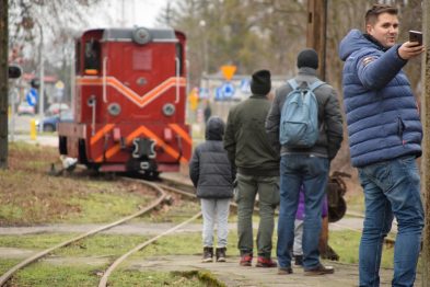 Grupa osób stoi na peronie przy torach kolejowych; w tle widać czerwony lokomotyw. Mężczyzna trzyma smartfon i robi zdjęcie, inni obserwują lokomotywę. Wokół jest posępny, zimowy dzień i ślady opadów na ziemi.