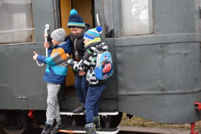 Dwoje dzieci w zimowych ubraniach stoi na stopniu starego pociągu, przygotowując się do wejścia do środka. Starsze dziecko trzyma w ręku metalową koronkę od zabytkowego sygnału kolejowego, a drugie patrzy w jego stronę. Oba wydają się być zaangażowane w wymianę zdań lub grę.