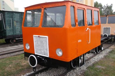 Pomarańczowy wózek motorowy stoi na torach kolejowych; posiada prostokątne reflektory i białe koła. Po bokach ma wyraźnie zaznaczone duże okna oraz białe drzwi. Na przedniej części wózka widoczny jest duży reflektor oraz srebrna kratka chłodnicy.