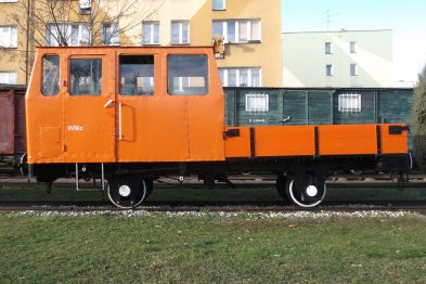 Pomarańczowy wózek motorowy stoi na torach obok zielonej trawy. Posiada dwa zestawy kół kolejowych, kabinę dla operatora i płaską platformę ładunkową. Budynki mieszkalne w tle sugerują, że wózek znajduje się w obszarze zurbanizowanym.