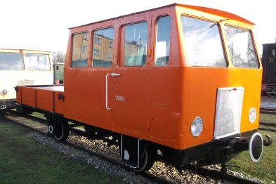 Odmalowany na pomarańczowo wózek motorowy stoi na torach kolejowych. Posiada kabiny z przeszklonymi oknami oraz platformę ładunkową. Wózek wyposażony jest w reflektory i zewnętrzne lusterka boczne.