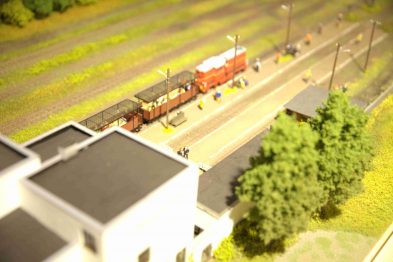 Miniaturka przedstawia fragment makiety kolejowej w skali H0 z lokomotywą parową i dwoma wagonami pasażerskimi. Model ukazuje torowisko otoczone zielenią z drzewami oraz mały budynek peronu. Makietę cechują dokładność wykonania i dbałość o detale realistycznie odwzorowujące środowisko kolejowe.