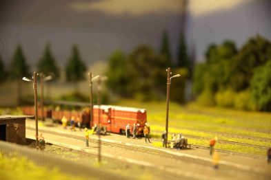 Model pociągu w skali H0 znajduje się na torach obok peronu przy którym widać postacie ludzi w miniaturze. Nad torami umieszczone są latarnie kolejowe, które oświetlają scenerię. W tle rozpościera się krajobraz z drzewami ustawionymi wzdłuż torów.