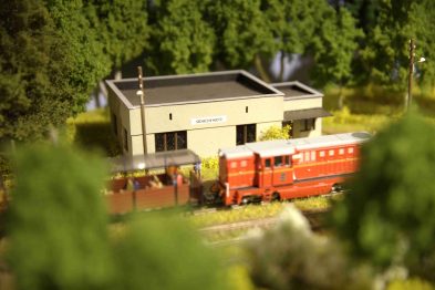 Czerwony model pociągu stoi na torach obok modelu niskiego budynku z napisem Sochaczew. Pociąg i budynek są otoczone drzewami oraz trawą, co nadaje scenie realistyczny wygląd miniaturowego krajobrazu. Scena jest częścią makiety kolei, która detalicznie odwzorowuje wygląd kolejnictwa i otoczenia.