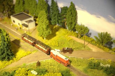 Model pociągu w skali H0 przemierza makietę krajobrazu, mijając przydrożne drzewa i niewielki budynek, ustawiony przy torach. Lokomotywa wraz z kilkoma wagonami toczą się po zakrzywionym odcinku torów. W otoczeniu makiety widać także naśladowane tereny zielone i skaliste, które nadają scenie realistyczny wygląd.