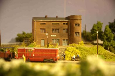 Model kolejowy przedstawia budynek z cegły z wieżą. Obok budynku znajduje się model czerwonego wagonu towarowego i figurka człowieka. Teren wokół jest udekorowany naśladując trawę i krzewy.