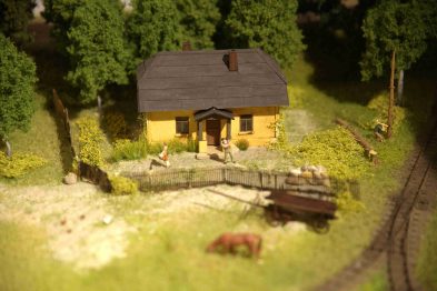 Makieta kolejowa H0 prezentuje żółty budynek z ciemnym dachem, otoczony zielenią i kwitnącymi krzewami. Przed domkiem znajdują się figurki ludzi, a tuż za drewnianym płotem widać model krowy. Tor kolejowy przebiega w pobliżu domu, podkreślając tematykę kolejową makiet.