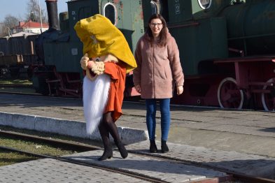 Osoba przebrana za postać z żółtą dużą głową i białą sukienką trzyma rękę na ramieniu kobiety ubranej w różowy płaszcz i czarne legginsy. Obydwie stoją na tle starych, zielonych lokomotyw parowych umieszczonych na torach kolejowych. Jest słoneczny dzień i obie postacie wydają się być w radosnym nastroju.
