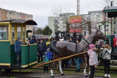 Na zdjęciu widoczna jest wąskotorowa lokomotywa i wagon, wokół których gromadzą się ludzie, w tym dzieci. W centrum stoi koń, który jest prowadzony przez osobę. Z tła wyłania się miejski krajobraz z budynkami mieszkalnymi.