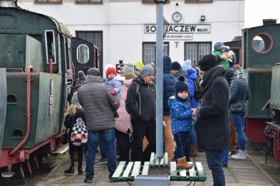Grupa osób, w tym dorosłych i dzieci, stoi na peronie wokół zielonej lokomotywy wąskotorowej i wagonów kolejowych. Niektórzy uczestnicy są ubrani w ciepłe zimowe ubrania jak czapki i kurtki. Widoczny jest również budynek z napisem 