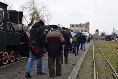 Grupa osób stoi na peronie obserwując wystawione eksponaty kolejowe; w tle widoczna jest parowa lokomotywa na torach. Uczestnicy są ubrani w zimowe odzienie, sugerujące chłodną pogodę. W otoczeniu peronu i torów znajduje się niska roślinność oraz zabudowania przemysłowe.