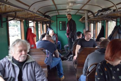 Wewnątrz wagonu kolejowego siedzą i stoją ludzie. Osoby zasiadają na drewnianych ławkach ustawionych wzdłuż ścian wagonu. W tle przez okno wagonu widać zewnętrze i światło dzienne.