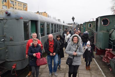 Grupa osób w różnym wieku spaceruje między starymi, zielonymi wagonami kolejowymi; niektórzy uśmiechają się do kamery. Chmurne niebo w tle i betonowe podłoże sugerują zimną pogodę. Na pierwszym planie widać kobietę z różową torebką trzymającą w dłoni białą parasolkę.