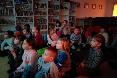Grupa dzieci siedzi na podłodze i skupiona ogląda coś przed sobą. W pomieszczeniu panuje półmrok, a ściany są wypełnione półkami z książkami. Dzieci są różnych wieków, a większość z nich ma na sobie kolorowe ubrania.