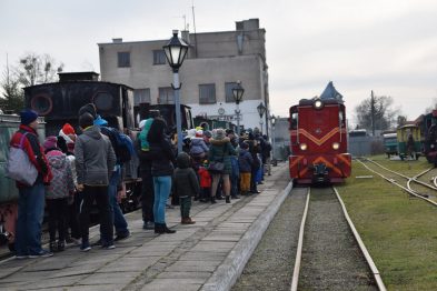 Grupa osób oczekuje na stacji kolejowej na pociąg wąskotorowy, który stoi na torach i jest gotowy do odjazdu. W tle widać budynek stacji i inne wagony kolejowe. Ludzie są ubrani w zimowe kurtki, co wskazuje na chłodną porę roku.
