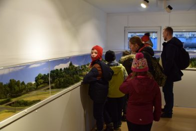 Grupa osób ogląda makiety kolejowe umieszczone wewnątrz wnętrza. Dzieci w kolorowych czapkach z zaciekawieniem przyglądają się mminiaturze krajobrazu z torami. Osoba dorosła towarzyszy dzieciom i wskazuje na jeden ze szczegółów ekspozycji.