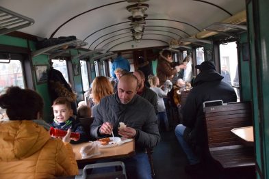 Wnętrze starego wagonu kolejowego pełne jest pasażerów; widać dorosłych i dzieci zajętych rozmową lub zajadających przekąski. Drewniane ławki zapewniają siedzenia dla podróżujących, a nad głowami wiszą lampy. Wśród pasażerów panuje miła, rodzinnie podkreślona atmosfera, a dzieci wydają się być w dobrych humorach.