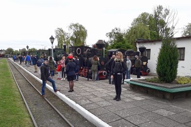 Grupa ludzi stoi na peronie przy torach kolejowych, gdzie zaparkowane są parowozy. Wśród zwiedzających widać osoby w różnym wieku, w tym rodziny z dziećmi. W tle znajdują się budynki muzeum oraz inne eksponaty kolejowe.