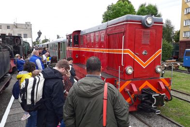 Grupa osób stoi przy czerwonym lokomotywie wąskotorowym, na którego boku widoczne są ozdobne żółte linie. Niektórzy pasażerowie wsiadają do pociągu, jeden mężczyzna prowadzi rower. Na tle pokrytych chmurami nieba, za lokomotywą dostrzec można inne maszyny kolejowe i zieleń drzew.