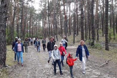 Grupa ludzi spaceruje leśną drogą porośniętą iglastymi drzewami. Większość osób nosi odzież odpowiednią dla chłodniejszej pogody, a niektórzy mają na sobie kurtki i plecaki. Widać także, że uczestnicy wycieczki rozmawiają między sobą podczas spaceru.