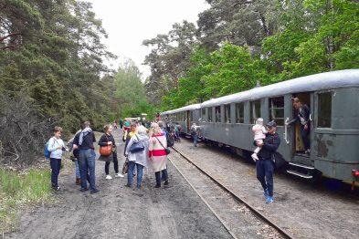 Ludzie wysiadają z szeregu starych, stalowych, zielonych wagonów kolejowych stojących na torach otoczonych drzewami. Niektórzy są ubrani w lekkie kurtki i trzymają torebki lub plecaki, wskazując na nieformalną atmosferę wycieczki. Wagon kolejowy ma otwarte drzwi, przez które pasażerowie schodzą na żwirową ścieżkę.