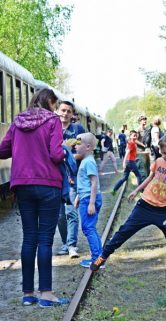 Starszy mężczyzna w ciemnym stroju, w tym kapeluszu, stoi obok starego pociągu, gdzie ludzie wchodzą i wychodzą z wagonów. Dzieci i dorośli mieszają się z obsługą na otwartej przestrzeni przy torze. Scena wydaje się być częścią większego wydarzenia lub ekskursji do historycznego składu kolejowego.