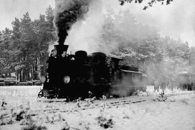 Czarno-biała fotografia przedstawia lokomotywę parową w ruchu na trasie otoczonej drzewami. Lokomotywa wydaje gęste kłęby dymu unoszące się do góry. Widoczne są również tory kolejowe oraz niewyraźne sylwetki osób obok pociągu.