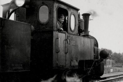 Stara parowa lokomotywa wąskotorowa jedzie po torach, emitując dym z komina. Maszynista jest widoczny w kabinie lokomotywy. Otoczenie jest nieostre, co sugeruje ruch pojazdu i stary charakter zdjęcia.