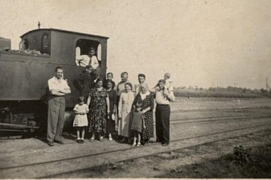 Lokomotywa wąskotorowa stoi na torach, obok której grupa osób pozuje do zdjęcia. Osoby są ubrane w odzież typową dla środkowego XX wieku. W tle widoczne jest otwarte pole oraz niewyraźna linia drzew.