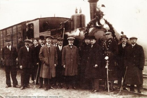 Grupa mężczyzn stoi przed parowozem i wagonem kolejowym; wszyscy są ubrani w stroje z epoki, które obejmują kapelusze i płaszcze. Na pierwszym planie widoczny jest mężczyzna trzymający laskę. Maszyna parowa ma klasyczną budowę z dużym kołem napędowym i jest wyposażona w reflektor na przedzie.