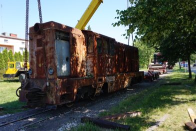 Stara lokomotywa spalinowa stoi na torach w dzień. Jest podnoszona przez dźwig, a w tle widoczne są drzewa i budynek. Lokomotywa ma rdzawą barwę i wiele oznak zużycia.