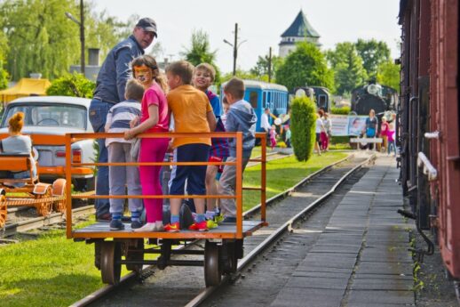 Grupa dzieci w towarzystwie dorosłego stoi na platformie wagonu kolejki wąskotorowej. W tle widoczne są zabytkowe wagony motorowe oraz zajęta peronem młodzież. Słońce świeci, a uczestnicy wydarzenia wydają się być w dobrych nastrojach.