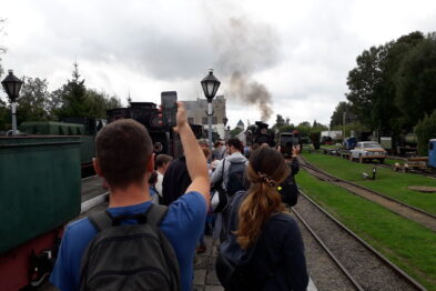 Grupa osób stoi na peronie kolejowym, niektóre z nich robią zdjęcia lub filmują. Parowóz wydobywa kłęby dymu na tle zadrzewionego terenu. W tle widoczna jest stacja kolejowa z zegarem.