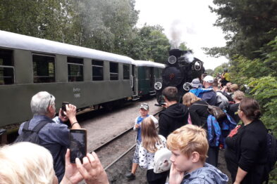Grupa osób stoi na peronie kolejowym, a niektóre robią zdjęcia i filmują przybywający parowóz ciągnący wagony pasażerskie. Parowóz wydaje dym, a tłum wydaje się zainteresowany tym zabytkowym pociągiem. Dzieci i dorośli obserwują i wskazują na lokomotywę, co świadczy o atrakcyjności wydarzenia.