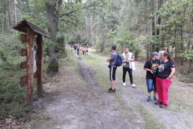 Grupa ludzi stoi na leśnej ścieżce obok drewnianej konstrukcji przypominającej małą wiatę lub kapliczkę. Trzy osoby przyglądają się małym przedmiotom, które jedna z nich trzyma w ręku. W tle za grupą widać innych uczestników spacerujących leśną drogą.
