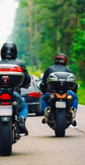 Dwóch motocyklistów jedzie drogą otoczoną drzewami, jeden ma czerwony motocykl, drugi czarny z dużymi bocznymi kuframi. W tle widać zieloną roślinność i delikatnie mglistą atmosferę. Nad nimi unosi się baner reklamowy z hasłem „Rajd Motocyklowy Industrialne Mazowsze”.