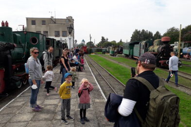 Ludzie stoją na peronie obok zielonej lokomotywy parowej i wagonów kolejki wąskotorowej. Dorośli i dzieci patrzą w kierunku torów, gdzie prawdopodobnie oczekują na nadjeżdżający pociąg. Scena odbywa się w dzień pod zachmurzonym niebem.