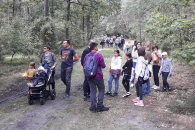 Grupa ludzi w różnym wieku spaceruje leśną ścieżką; niektórzy uczestnicy rozmawiają ze sobą, tworząc swobodną atmosferę. Osoba w tle prowadzi wózek z dzieckiem, podczas gdy inne dzieci i dorośli cieszą się wspólnym czasem. Drzewa tworzą zielone tło dla sceny, a uczestnicy wydarzenia wydają się mieć na sobie swobodny strój odpowiedni na spacer po lesie.