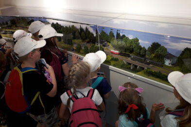Grupa dzieci stoi przy dużym oknie, przez które obserwują krajobraz z pojeździec kolejowym. Wszystkie dzieci mają na głowach białe kapelusze i plecaki, a na dworze widać zielone tereny i jasnoniebieskie niebo. Widoczna część modelu kolejowego z miniaturkami pociągów i szynami przyciąga uwagę młodych obserwatorów.