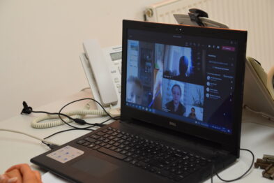Laptop stoi na biurku, a na ekranie wyświetlona jest aplikacja do wideokonferencji z kilkoma uczestnikami. Obok komputera leżą różne przedmioty takie jak telefon, długopis i notatnik. W tle widać tablicę ogłoszeń i inny sprzęt biurowy.