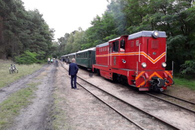 Czerwono-zielony pociąg retro stoi na torach w otoczeniu drzew. Po lewej stronie widoczna jest postać ludzka skierowana w stronę lokomotywy. Na drugim planie pojawiają się pasażerowie oraz rowerzyści, co świadczy o turystycznej atmosferze wydarzenia.