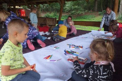 Grupa dzieci siedzi przy długim białym stole usytuowanym pod zadaszeniem i angażuje się w działania plastyczne, używając kolorowych kred i innych materiałów pisarskich. Dorośli nadzorują i wspomagają zajęcia, współpracując z młodszym pokoleniem. Sceneria jest leśna i rekreacyjna, zapewniając przyjemną atmosferę dla rodzinnych aktywności.