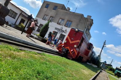 Czerwona lokomotywa wąskotorowa stoi na torach przed budynkiem z napisem 