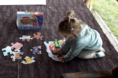 Dziewczynka siedzi na brunatnym dywanie składając puzzle, których tematyka również jest związana z pociągami. Część puzzli jest już ułożona, a także widoczne jest pudełko z obrazkiem złożonej układanki. Wokół dziecka rozsypane są pojedyncze elementy puzzli.