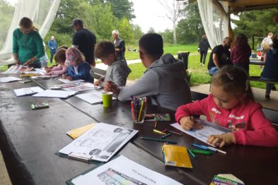 Grupa dzieci siedzi przy długim stole na zewnątrz i zajmuje się kolorowaniem i pisaniem. Na stole rozłożone są materiały plastyczne takie jak kredki i kartki z wizerunkami pociągów. Wokół stołu widać opiekunów i inne osoby uczestniczące w wydarzeniu.