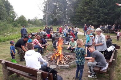 Grupa ludzi, w tym dorosłych i dzieci, gromadzi się wokół ogniska na zewnątrz; dzieci stoją najbliżej ognia, trzymając patyki. W tle widać drzewa i nieco dymu, a uczestnicy imprezy korzystają z ławek ustawionych wokół ogniska. Atmosfera wydaje się swobodna i przyjazna rodzinnej zabawie.