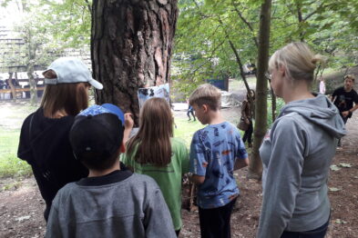 Grupa osób, w tym dzieci i dorośli, stoi przed drzewem rozglądając się i coś obserwując. Jedno dziecko trzyma w ręku kartkę papieru. W tle widoczne są inne osoby oraz elementy zielonego parku lub lasu.